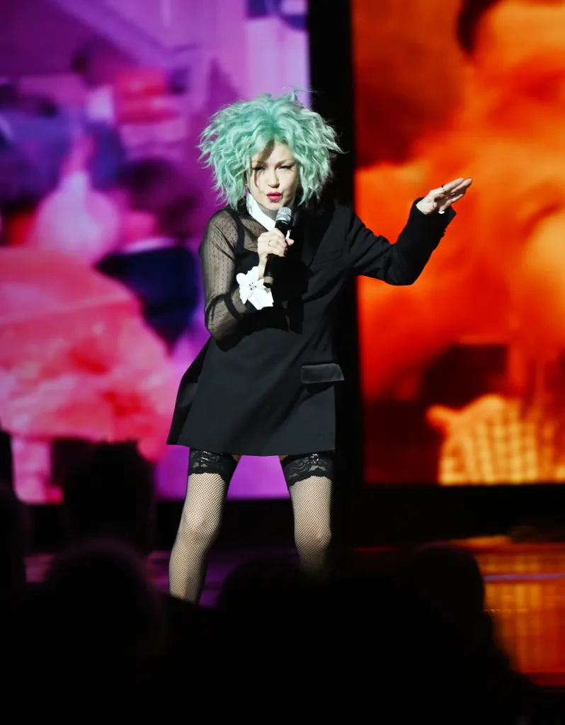 Cyndi Lauper performing live with a microphone, wearing a black blazer over a sheer top and fishnet stockings, with teal curly hair.