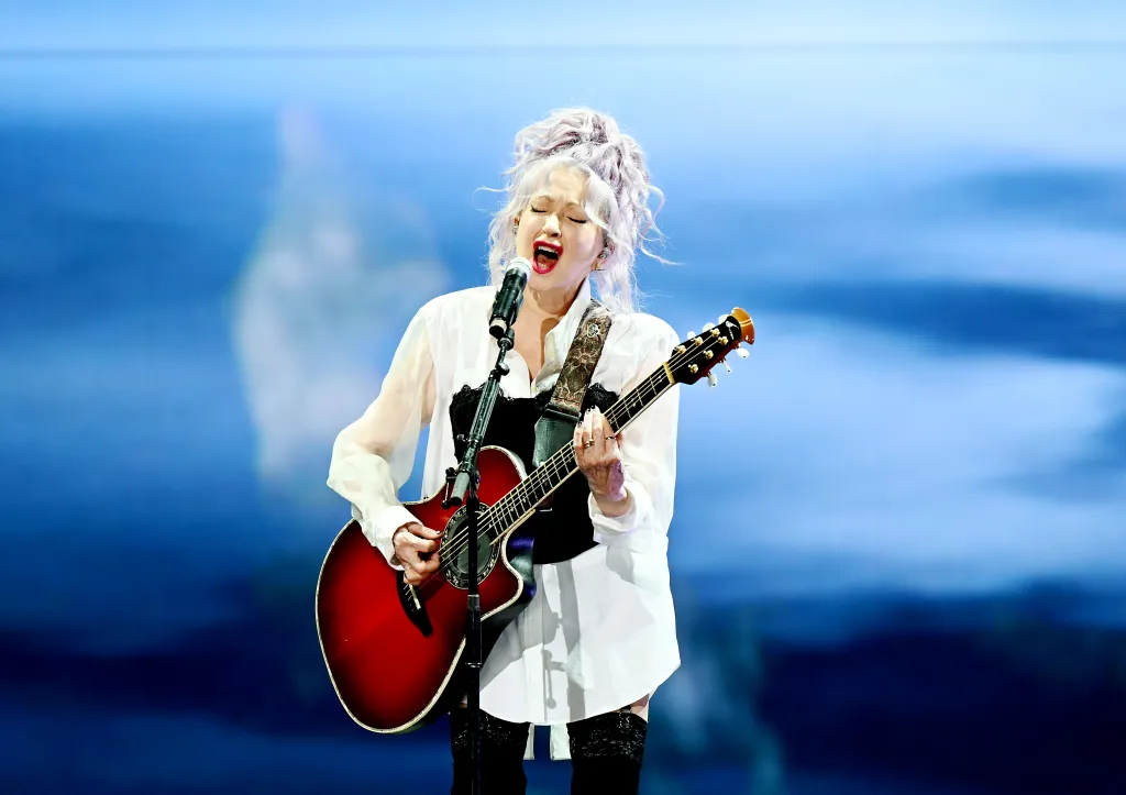 Cyndi Lauper performing live, singing into a microphone and playing an acoustic guitar.