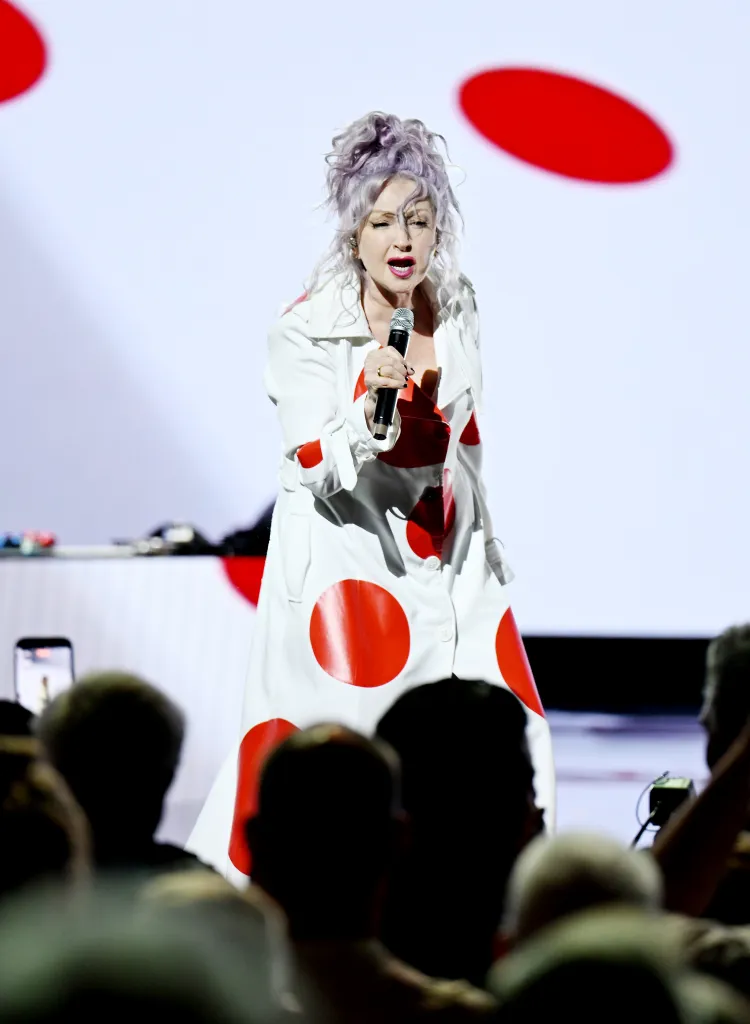 Cyndi Lauper performing in a white coat with large red polka dots, holding a microphone.