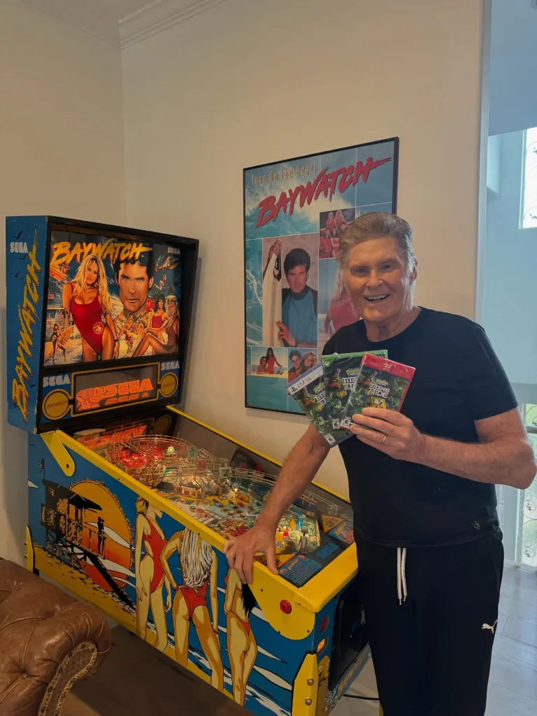 David Hasselhoff stands next to a Baywatch pinball machine and poster, holding three 