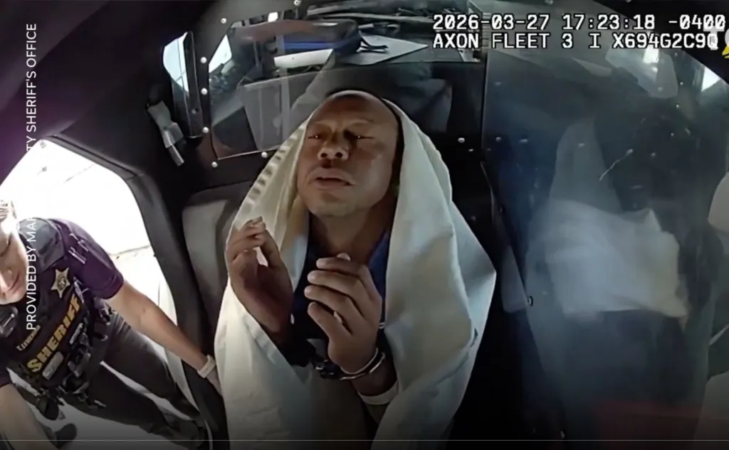 Tiger Woods in a police car, appearing dazed and wrapped in a white blanket, with a sheriff's deputy visible beside him.
