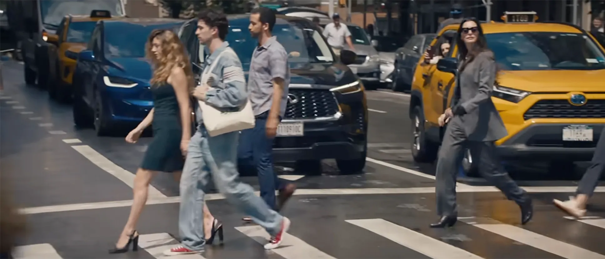 A street scene with people crossing the road, a woman in a black dress walks next to a man in jeans and a light jacket, both in front of a blue car. To their right, a woman in a grey pantsuit walks in front of a yellow taxi, next to another person.