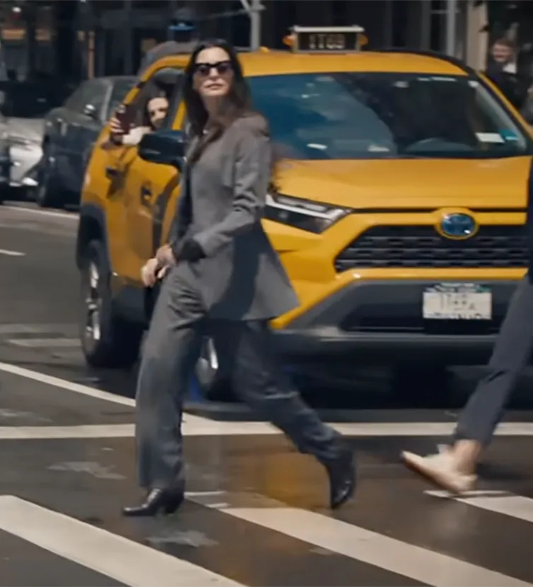 A woman in a gray suit crosses the street as someone in a yellow taxi films her.