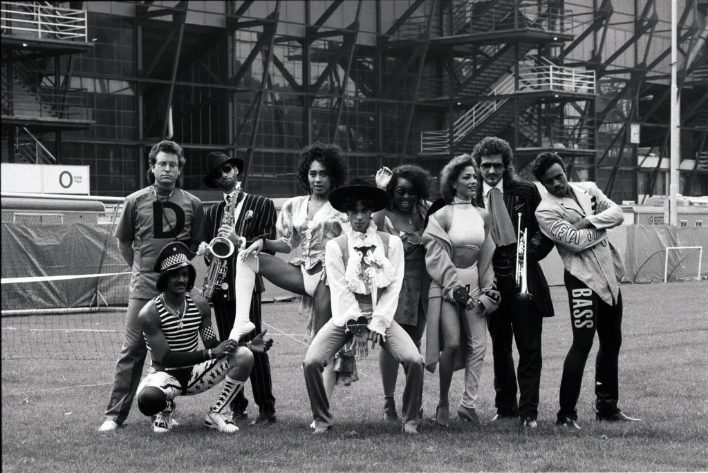 Prince and his band, including Dr. Fink, Bobby Z, Rosie Gaines, Boni Boyer, Sheila E, and Brownmark, posing on a grassy field.