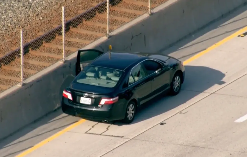 A dark sedan stopped on the shoulder of a highway, with the driver's side door open.