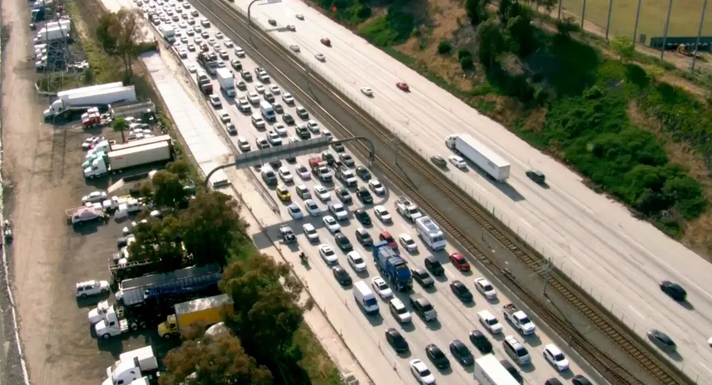 An aerial view of the 105 Freeway in South L.A. with heavy traffic in one direction and light traffic in the other, next to a parking area filled with trucks and cars.