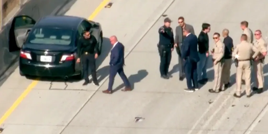 Law enforcement officers and civilians gather around a black sedan with its driver's side door open on a freeway.