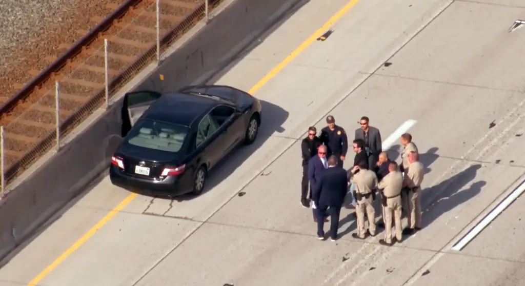 Overhead view of a black car on the side of a highway with its door open, and a group of police officers and men in suits standing near it.