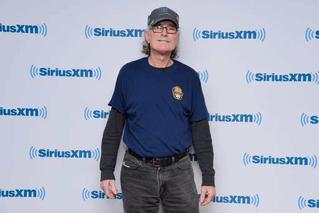 Drummer Vini 'Mad Dog' Lopez posing in a blue shirt with a badge, black jeans, and a cap.