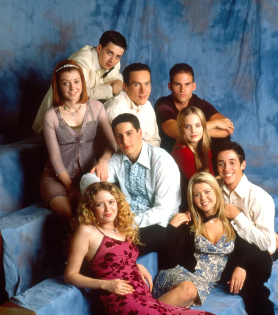 From left: Eddie Kaye Thomas, in a cream sweater, Chris Klein, in a white shirt, Seann William Scott, in a red sweater, Mena Suvari, in a red top, Thomas Ian Nicholas, in a white shirt, Tara Reid, in a blue printed dress, and black cardigan, Natasha Lyonne, in a pink patterned dress, Jason Biggs, in a frilled blue shirt, Alyson Hanniganin a brown dress and cardigan.