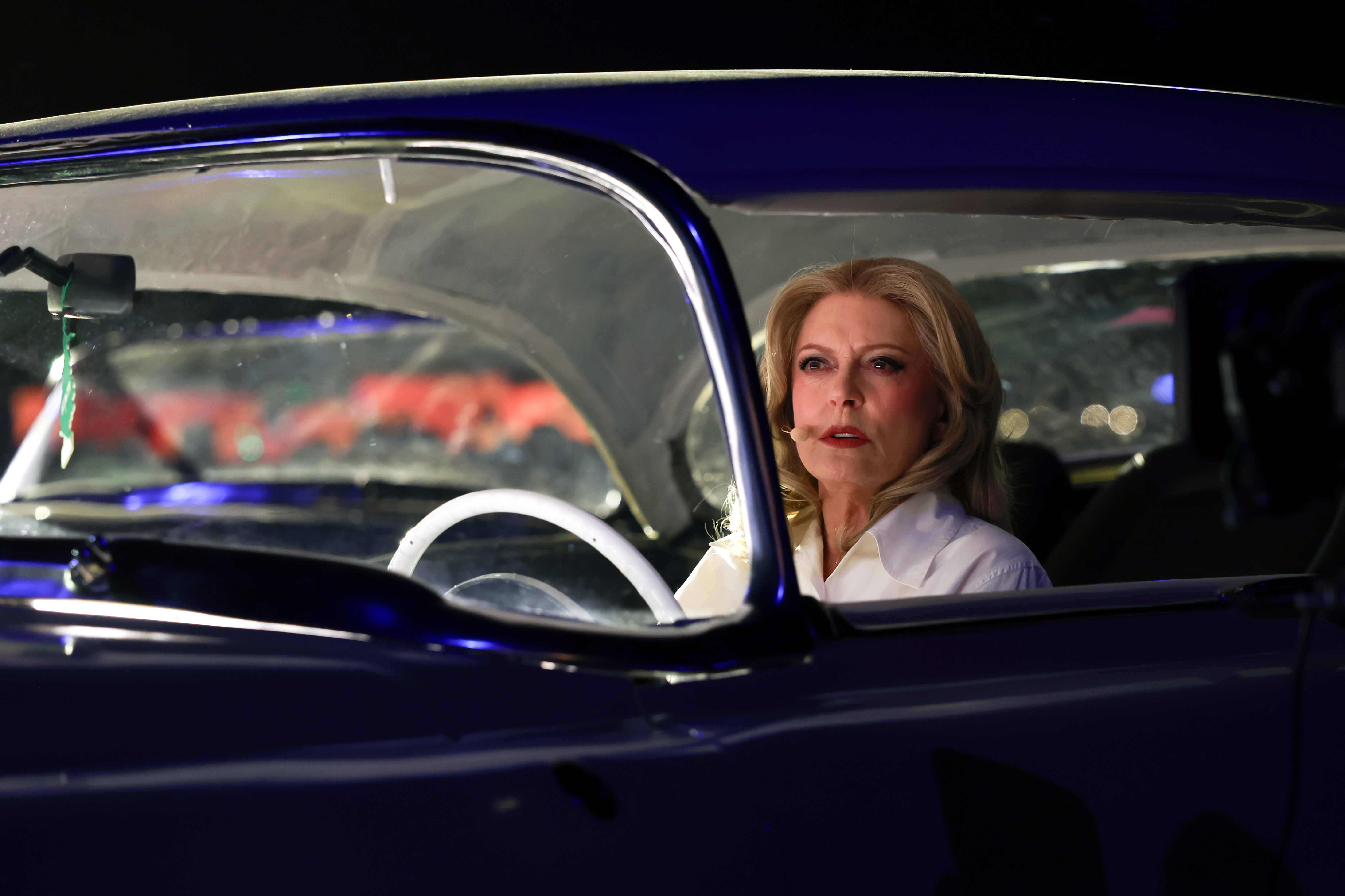Susan Sarandon in a white shirt, with blonde hair and red lipstick, sits behind the white steering wheel of a dark car, looking forward.