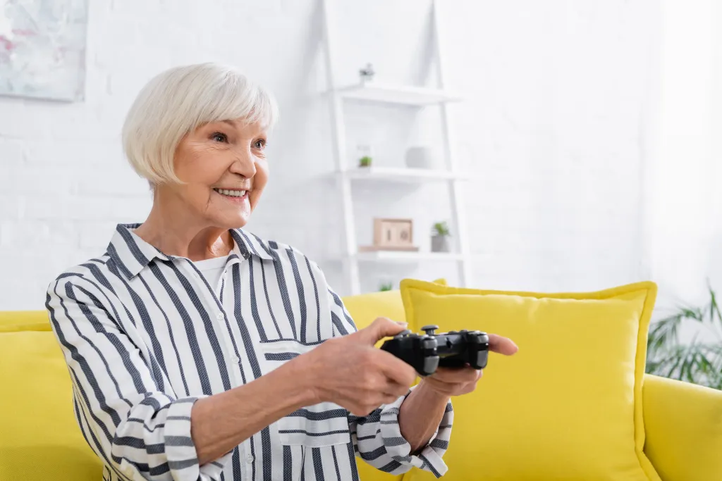 Elderly woman in striped shirt sitting on a yellow sofa and smiling while playing a video game.