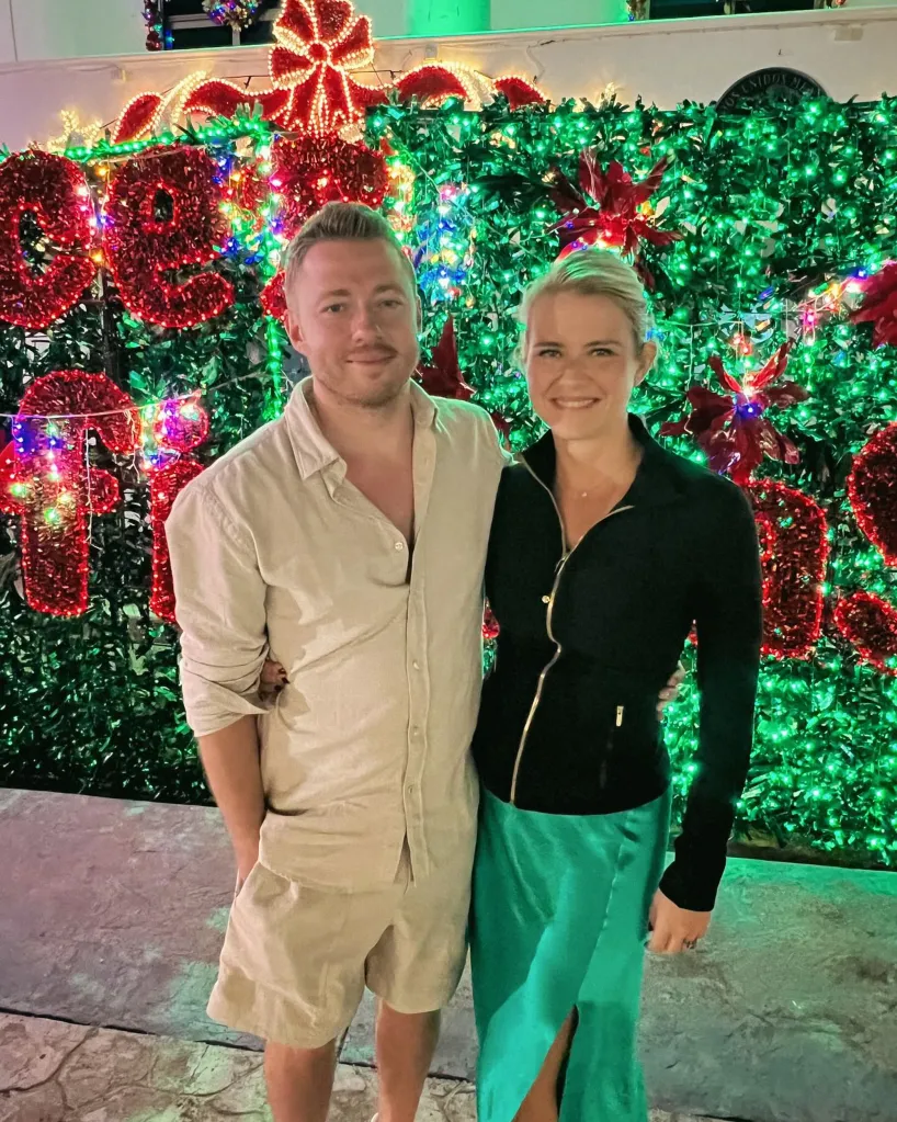 A man and a woman stand smiling in front of a wall of green foliage with holiday lights.