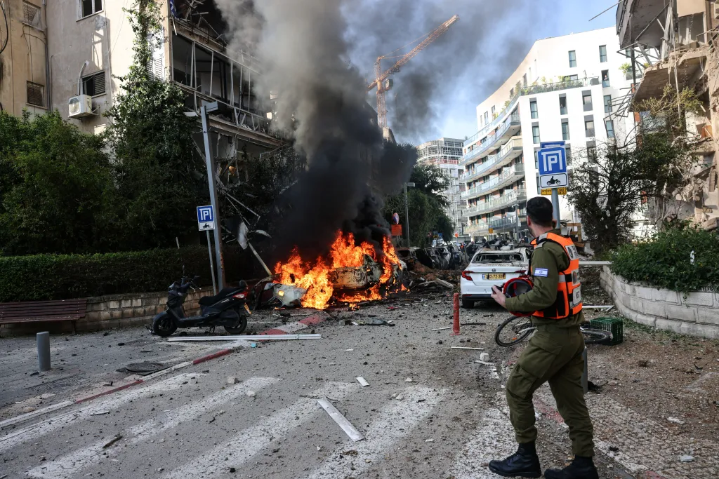 An emergency worker stands on a debris-filled street in front of a burning car and damaged buildings.