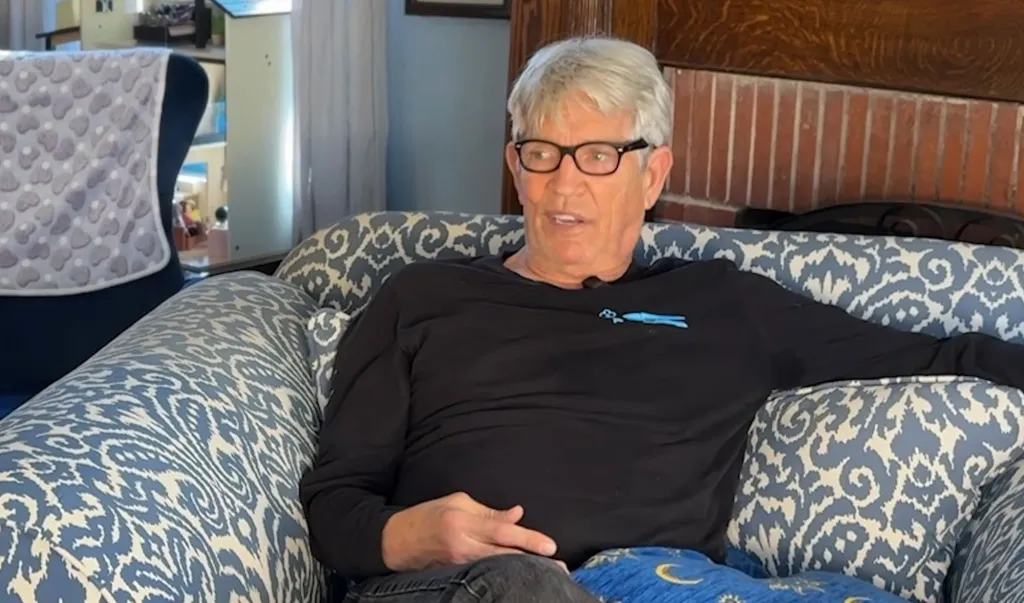 Eric Roberts sits on a patterned couch, speaking emotionally.