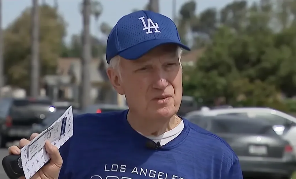 An elderly man in a blue Dodgers hat and shirt holds paper tickets.