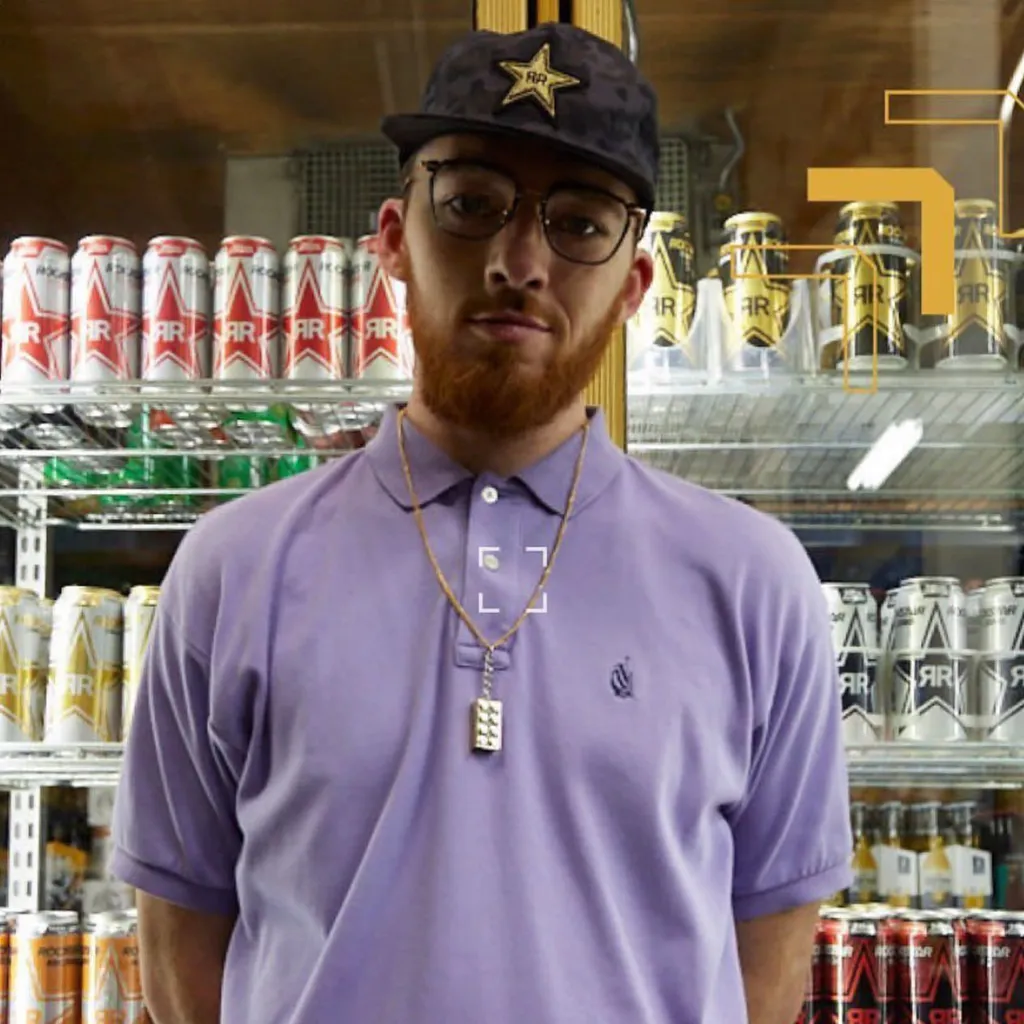 Angus Cloud, in a purple polo shirt and backward baseball cap, stands in front of a refrigerator stocked with Rockstar energy drinks.