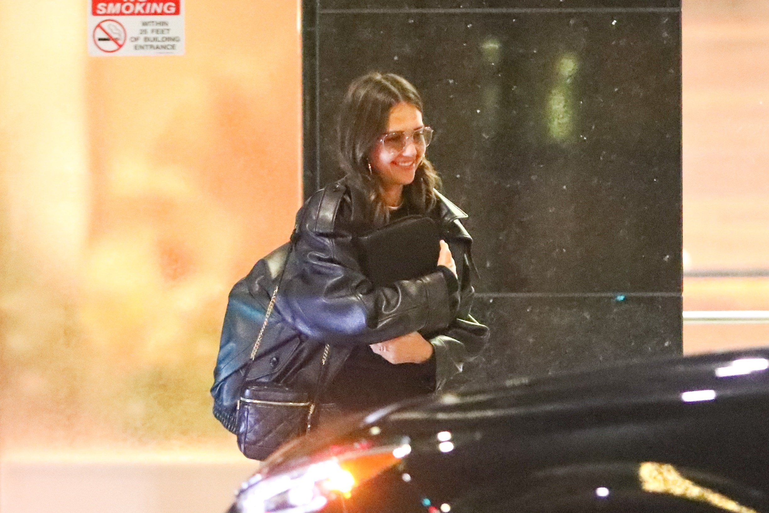 Jessica Alba smiling while wearing a black leather jacket, glasses, and carrying a quilted bag.