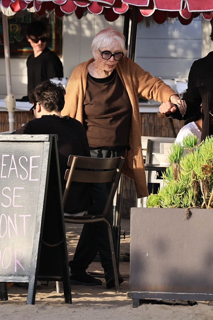 Shirley MacLaine in a brown shirt and cardigan, wearing glasses, holding onto an arm for assistance.