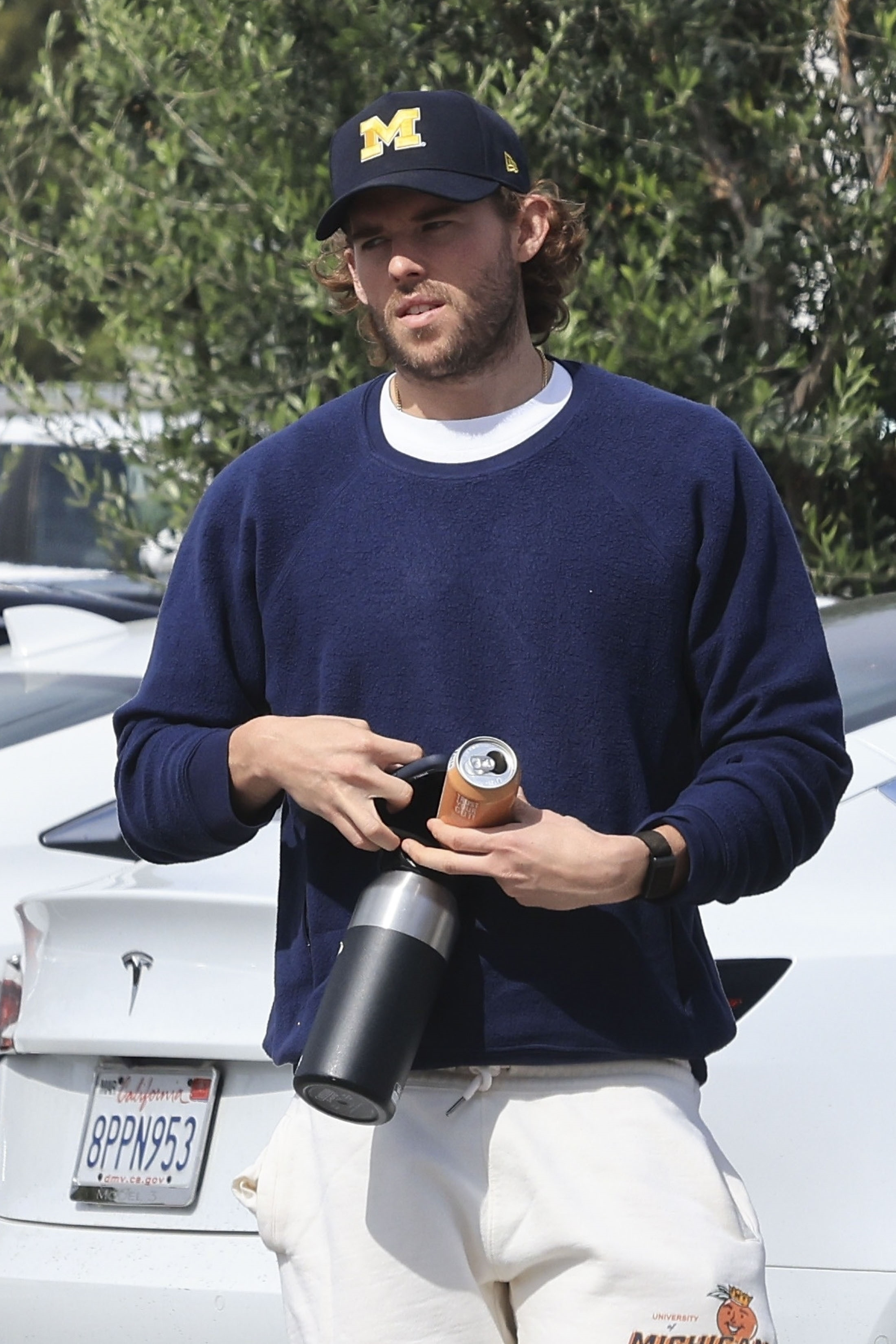 Christopher Schwarzenegger wearing a navy sweatshirt, off-white joggers, and a baseball cap, holding a drink can and a thermos.