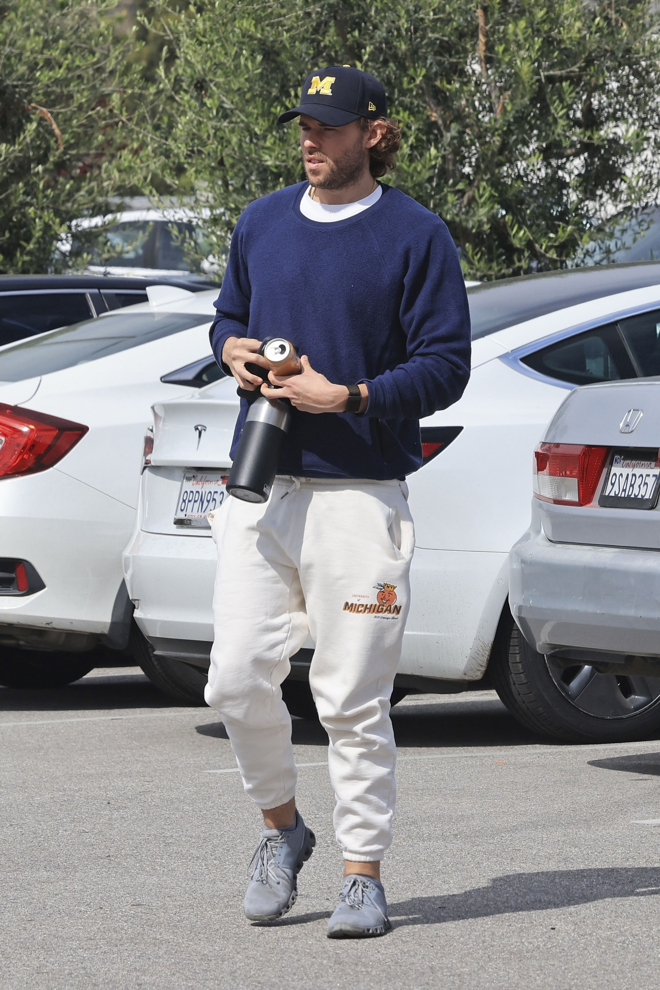 Christopher Schwarzenegger in a navy crewneck, off-white joggers, and a baseball cap, holding a drink.