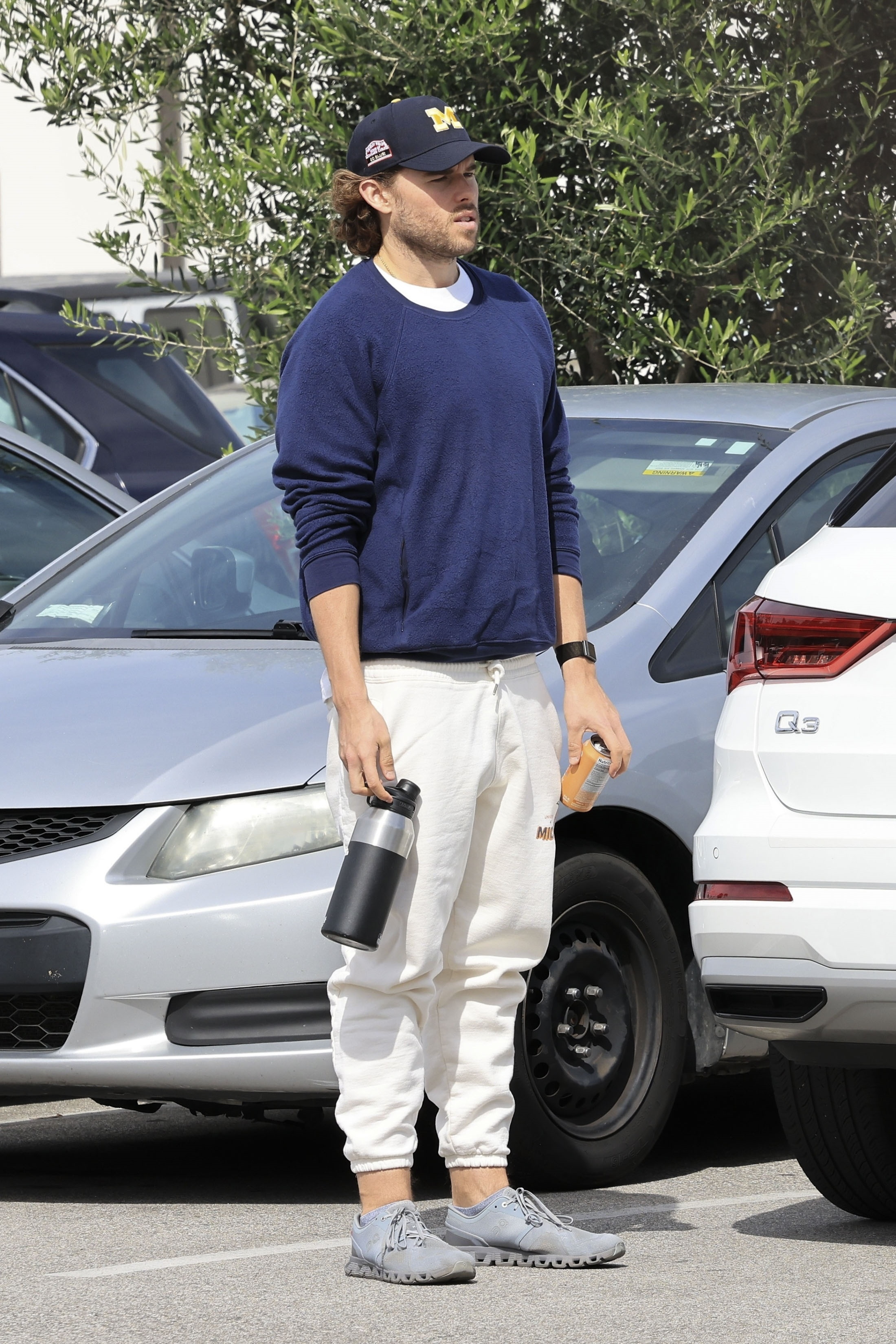 Christopher Schwarzenegger in Malibu wearing a navy crewneck sweatshirt, off-white joggers, a baseball cap, and sneakers, holding a water bottle and a can.