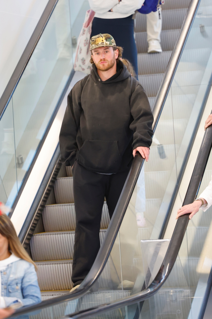 West Wilson standing on an escalator, dressed in a black hoodie, black pants, and a camouflage baseball cap, with other people around him.