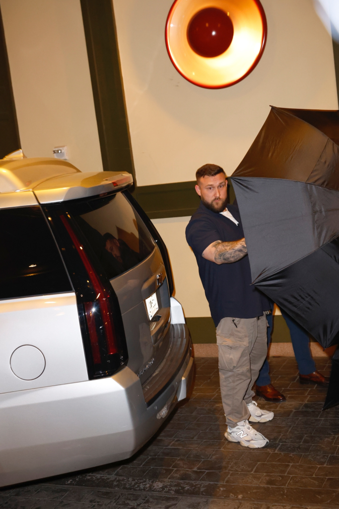 A man with a beard and tattoos holds open a large black umbrella, partially obscuring the view of Taylor Swift and Travis Kelce as they arrive at Casa Cipriani in NYC.