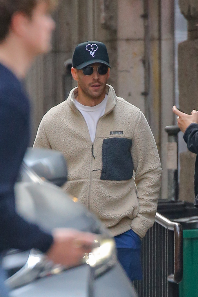 Kyle Cooke, a reality star, walks in a New York City street wearing a cream fleece, blue shorts, a black hat with an elephant logo, and sunglasses.