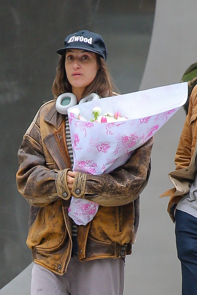Amanda Batula walking down the street in a distressed leather jacket, baseball cap, and headphones, carrying a bouquet of flowers.