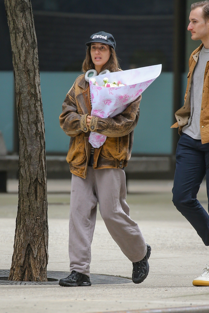 Amanda Batula in a brown leather jacket, grey sweatpants, and a black baseball cap, holding a bouquet of flowers, walking next to a man.