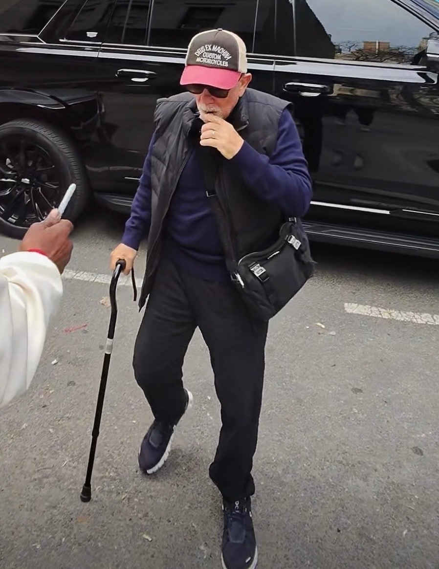 Billy Joel in incognito wearing a hat, sunglasses, vest, and carrying a cane.