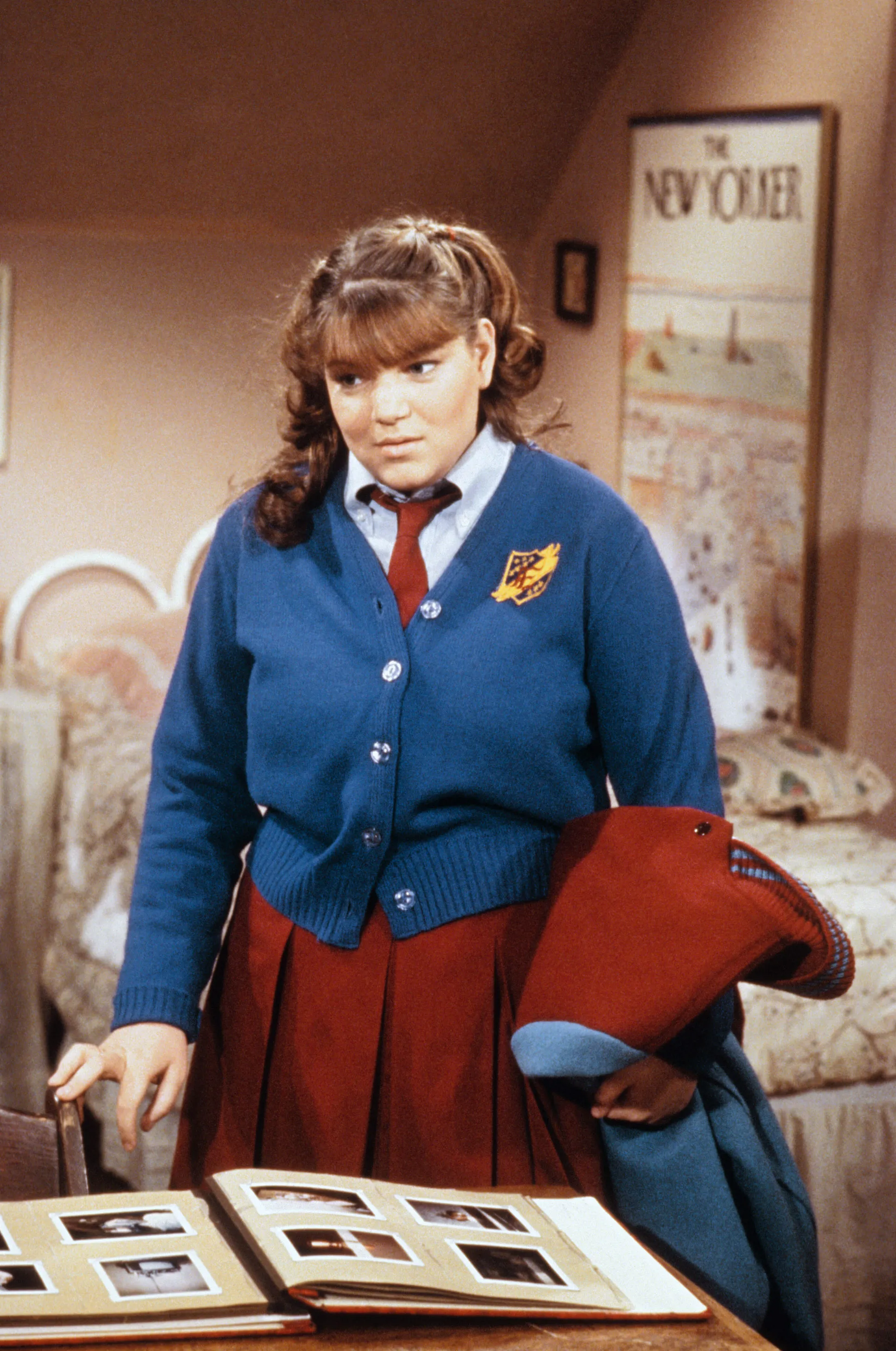 Mindy Cohn in a school uniform, holding a coat over her arm, looking at photo albums.