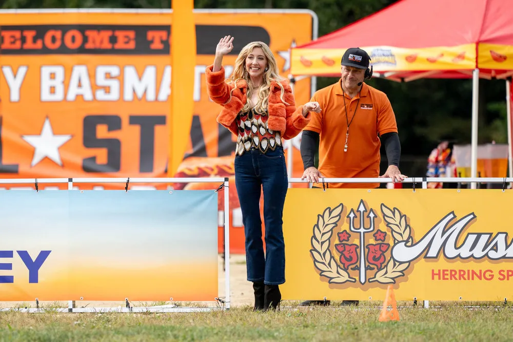 Heidi Gardner as Tisha Basmati waves next to a man in an orange shirt at an outdoor event.