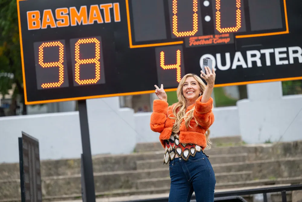 Heidi Gardner as Tisha Basmati taking a selfie in front of a scoreboard.