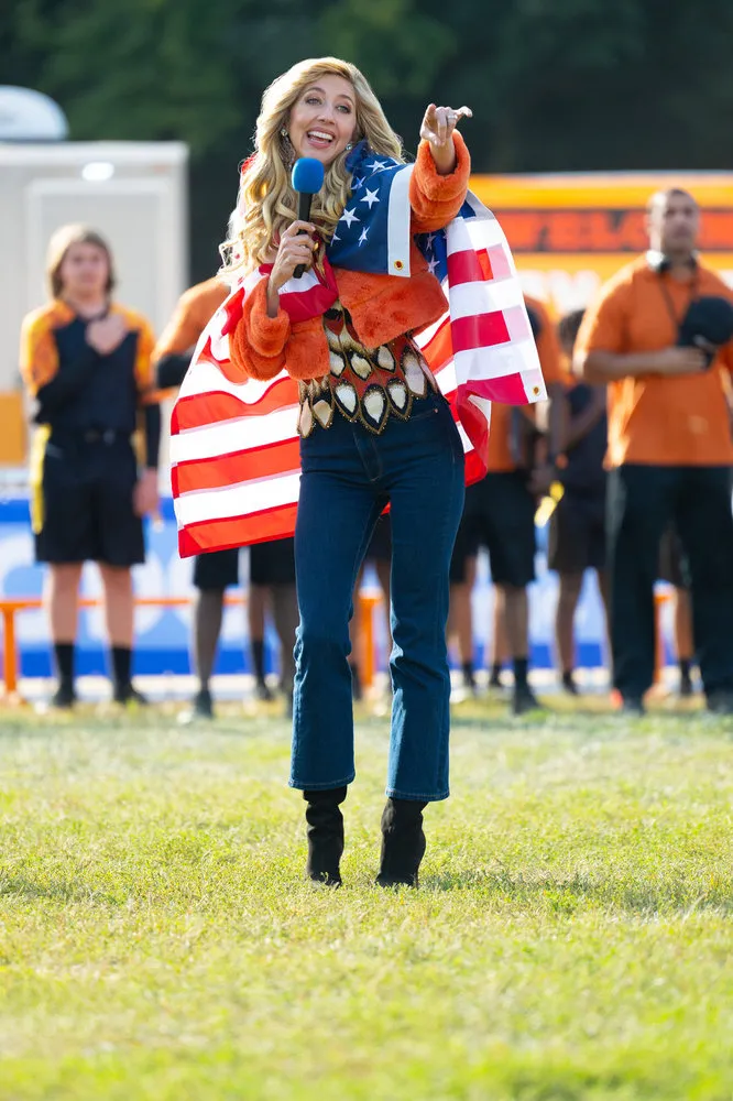 Heidi Gardner as Tisha Basmati, wearing an orange jacket, an American flag, and jeans, holding a microphone and pointing.