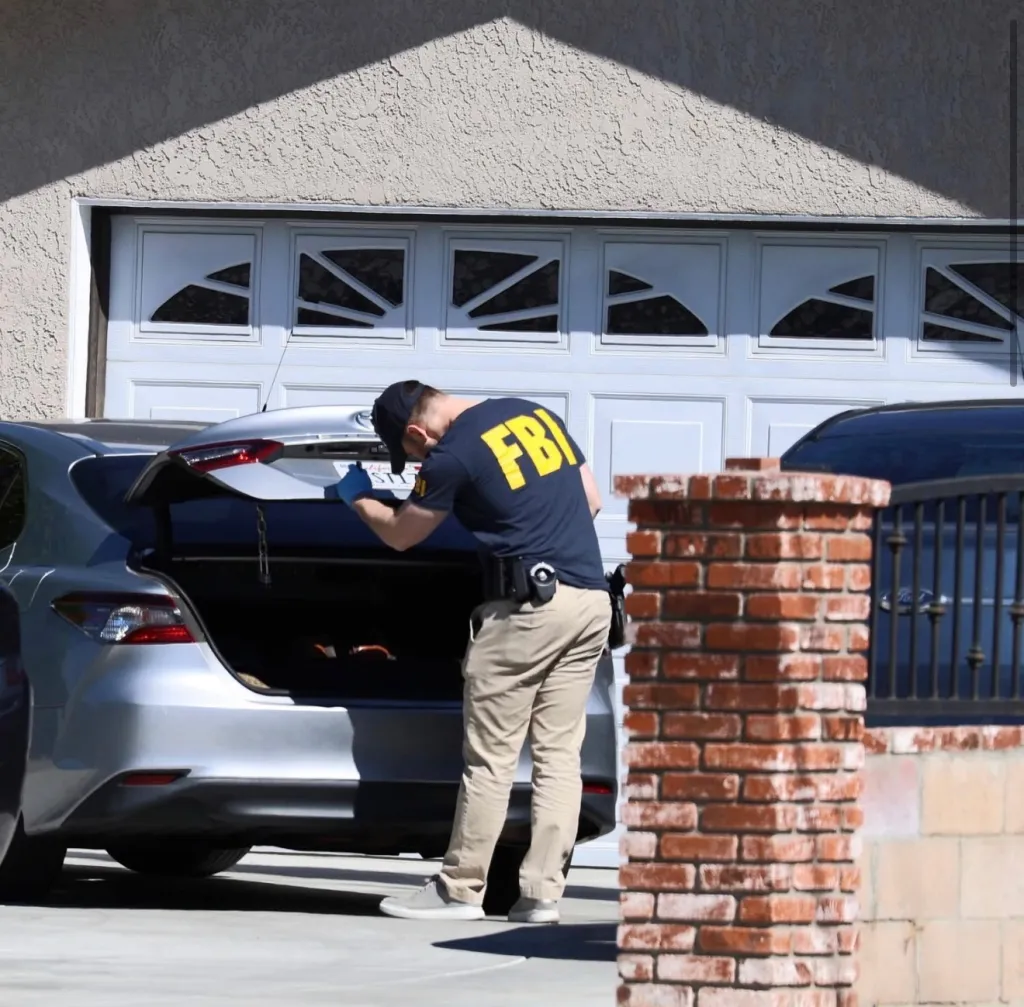 FBI agent in a T-shirt and khakis inspecting the trunk of a silver car parked in a driveway.