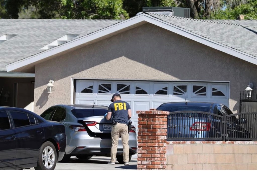 An FBI agent standing behind a silver car in a driveway in front of a house.