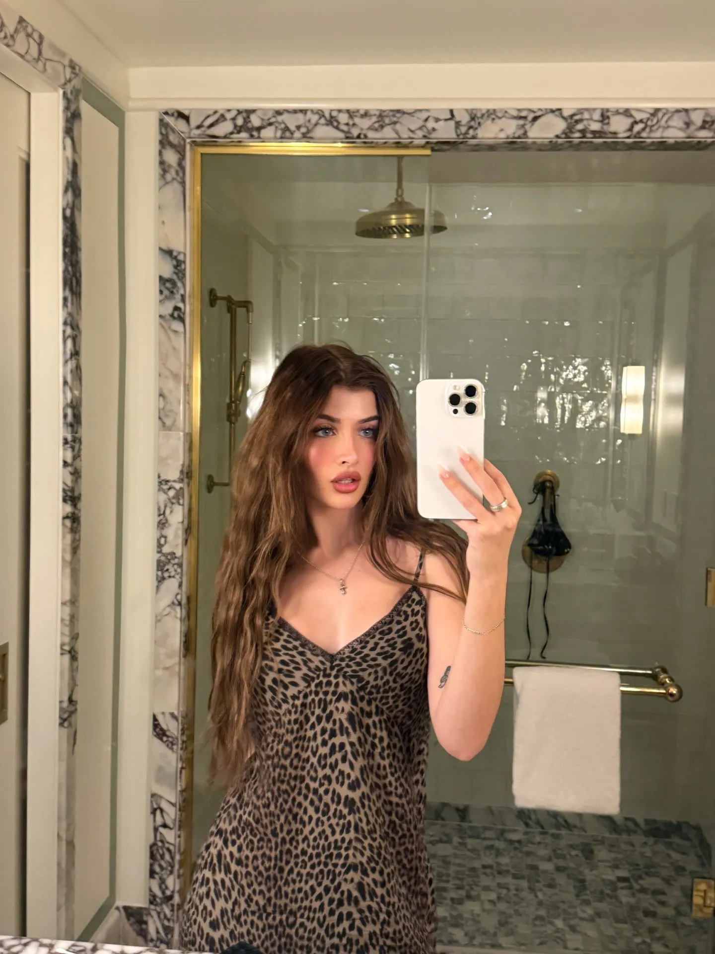 A woman with long wavy brown hair, wearing a leopard print dress, takes a mirror selfie in a bathroom with marble accents.
