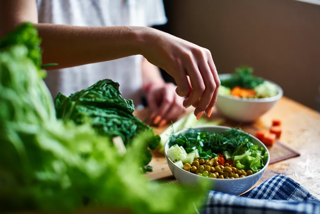 Female hand pouring green onions into a bowl of salad with peas, cucumbers, carrots, lettuce, and dill.