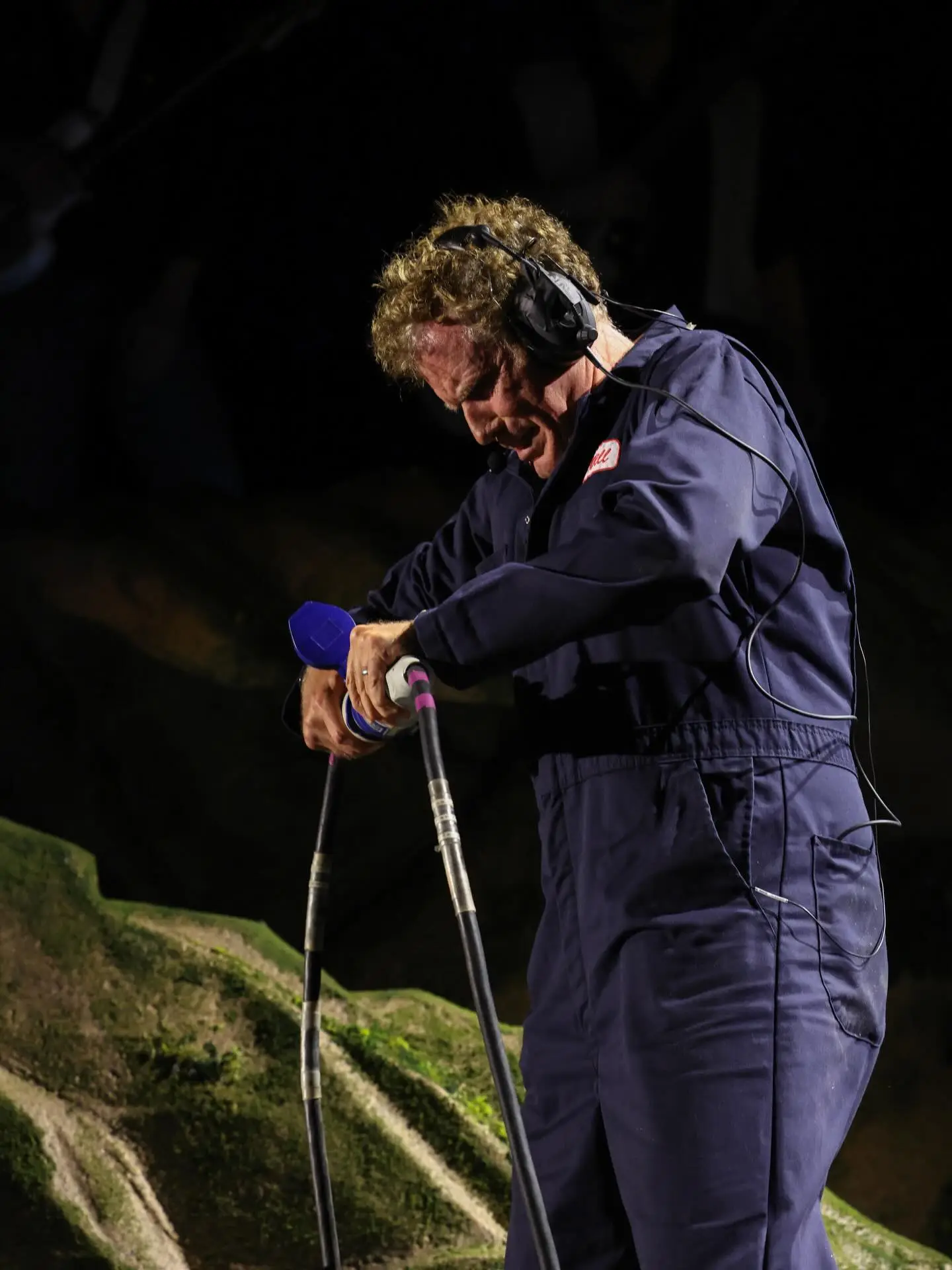 Will Ferrell in a blue jumpsuit with headphones holding a prop microphone during a performance.