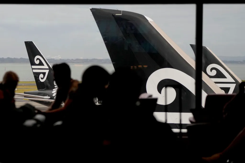 Passengers in a lounge at Auckland International Airport view two Air New Zealand planes through a window.
