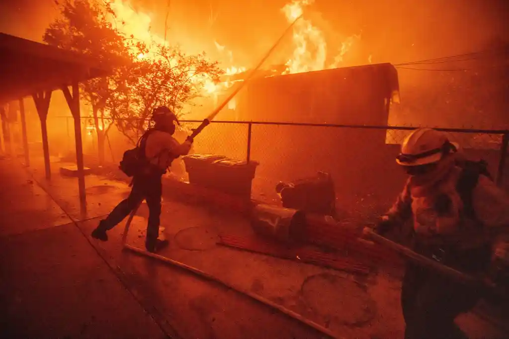Two firefighters battling a blaze as a structure burns in the background.