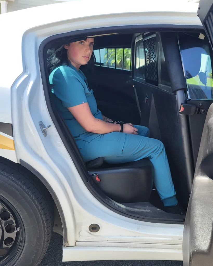 Woman in teal scrubs sitting in the back of a police car.