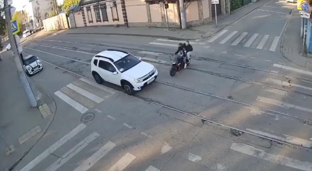 Footage of a motorcycle colliding with a car at an intersection, throwing the rider and passenger onto the roadway.