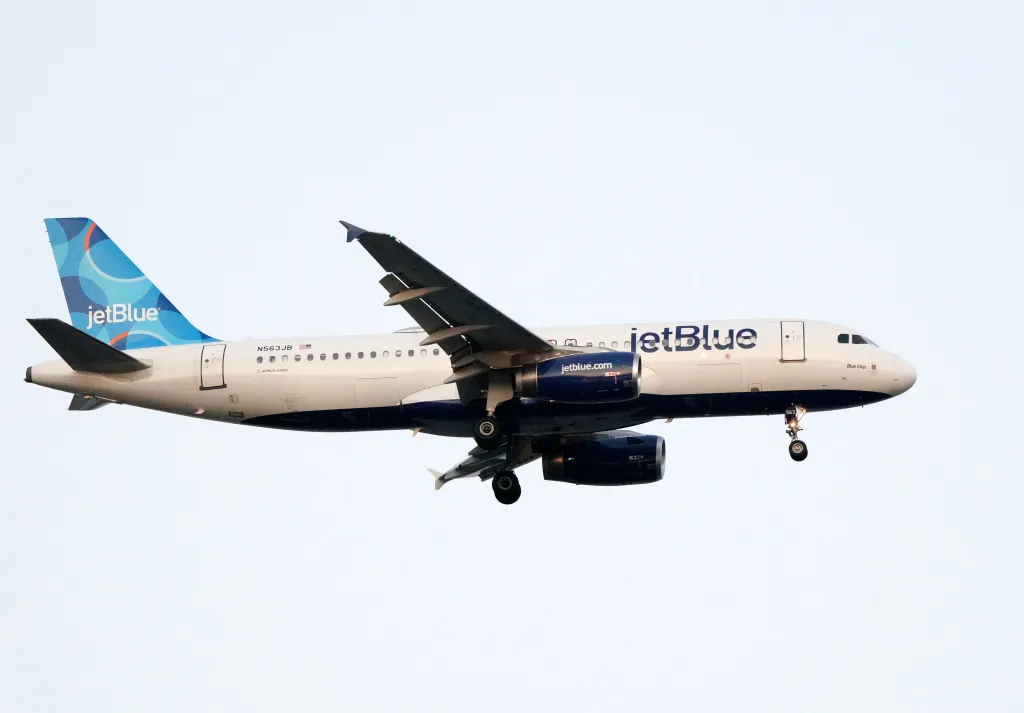 A white JetBlue Airbus A320 airplane with blue engines and tail, and landing gear extended, flies across a white sky.
