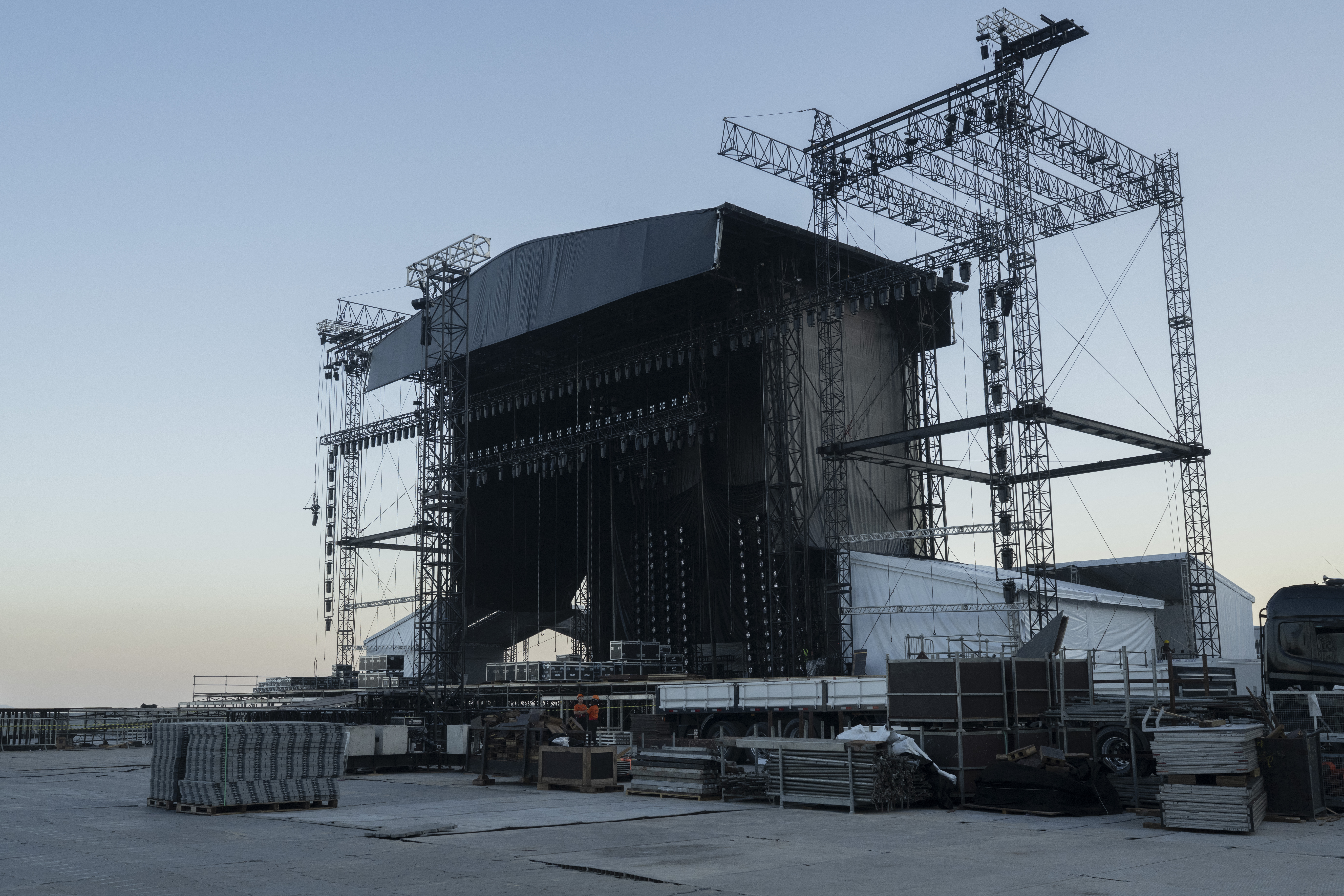 A large, partially constructed stage with metal scaffolding and black fabric, surrounded by equipment, after a worker died during its setup.