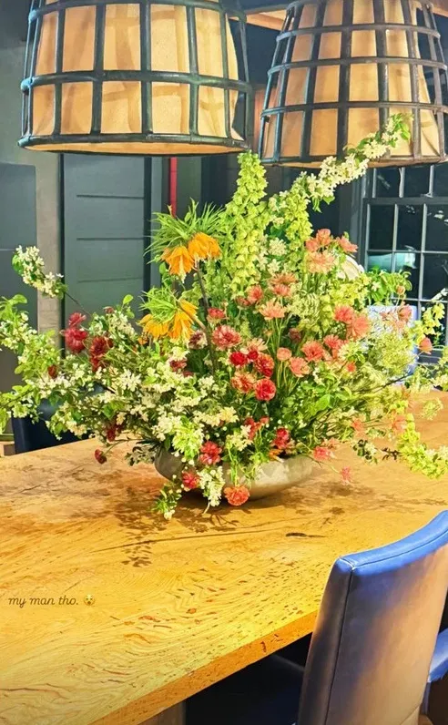 A large floral arrangement with a variety of red, orange, white, and green flowers on a wooden table, with large basket-like light fixtures overhead.