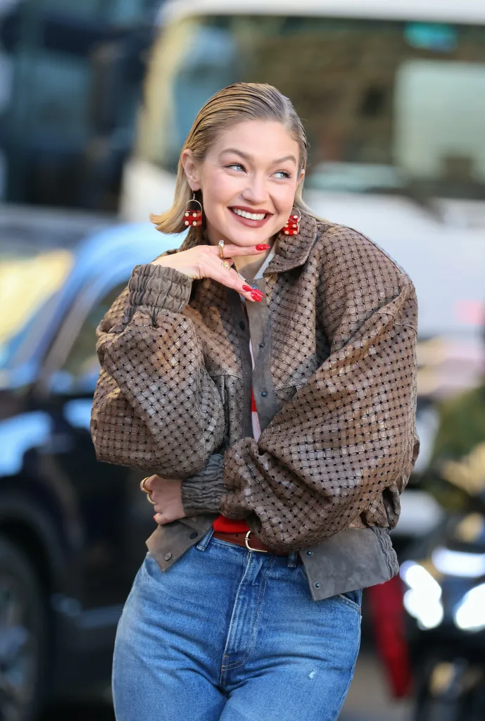 Gigi Hadid filming a commercial, wearing a brown textured jacket, blue jeans, and red dice earrings, smiling with her hand on her chin.