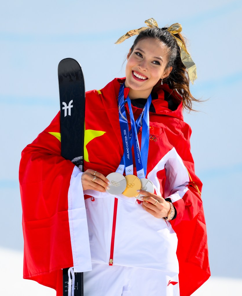 Ailing Eileen Gu holding three medals and wrapped in the Chinese flag.